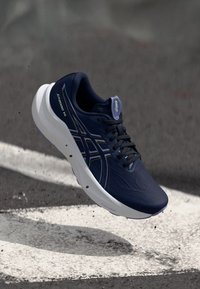 Marineblauer Laufschuh mit einer glatten, strukturierten Oberfläche, ausgestattet mit Schnürsenkeln und einem Logo, sowie einer weißen gepolsterten Sohle mit dunklen Akzenten.