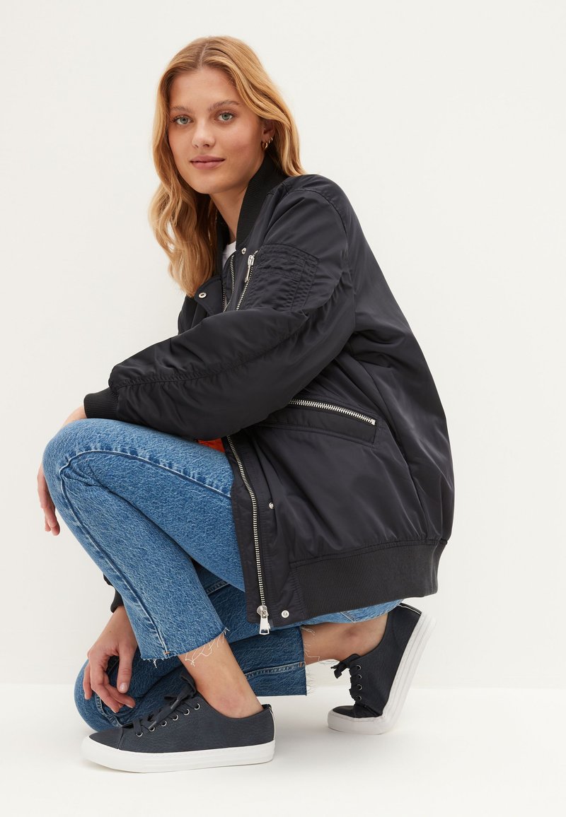 Melna bombu jaka ar izšūtiem piedurknēm, kabatām ar rāvējslēdzēju un ribotām aprocēm, kombinēta ar nodilušām zilajām džinsām un tumši zilām sporta kurpēm.