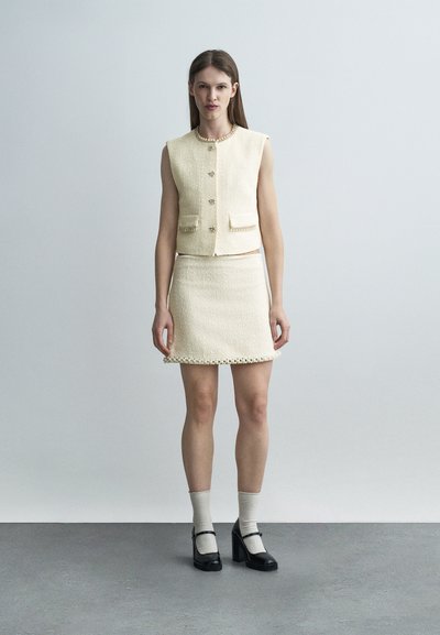 Γυναίκα που φοράει κρεμ αμάνικο υφασμάτινο γιλέκο και ταιριαστό φούστα, μαύρες γόβες Mary Jane με ανοιχτόχρωμες κάλτσες, στέκεται σε γκρι πάτωμα, απλό φόντο.