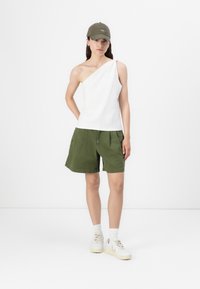 Top alb, cu un umăr, cu detaliu nod, asortat cu pantaloni scurți de un verde măsliniu. Adidași albi cu accente bej. O șapcă neutră completează ținuta.