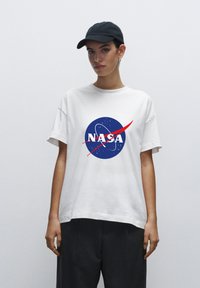 Bílý bavlněný tričko s modrým logem NASA a červenými akcenty. Model má na sobě černou kšiltovku, ukazuje uvolněný, oversized střih.