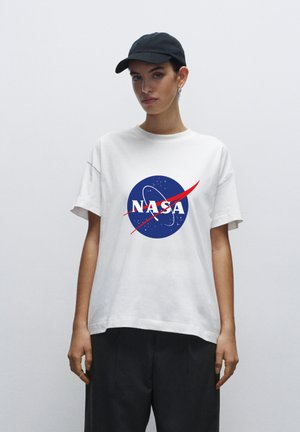 Hvid bomulds T-shirt med et blåt NASA-logo og røde detaljer. Modellen bærer en sort kasket og viser en afslappet, oversized pasform.