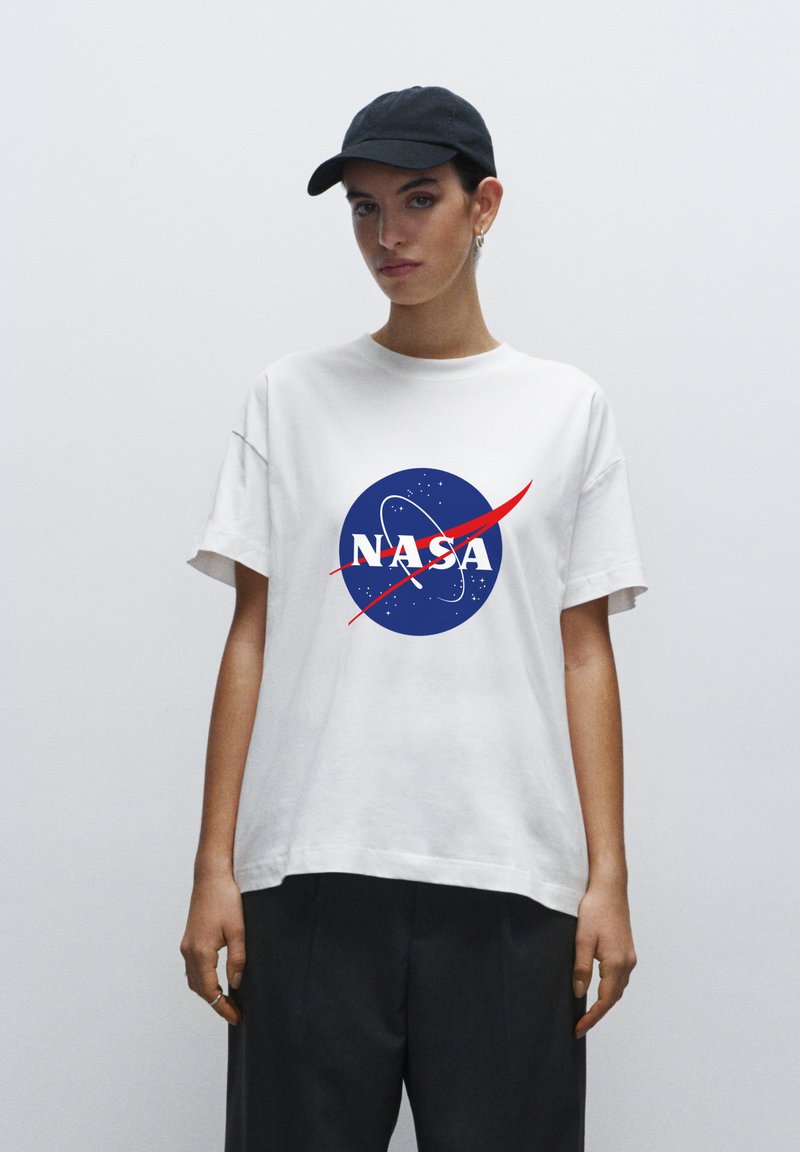 Bílý bavlněný tričko s modrým logem NASA a červenými akcenty. Model má na sobě černou kšiltovku, ukazuje uvolněný, oversized střih.