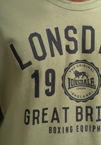 Olajzöld pamut póló, amelyen merész fekete szöveg olvasható: "LONSDALE 19 NAGY-BRITANNIA BOXING FELSZERELÉS", és egy kör alakú logó látható, rajta egy oroszlánnal.