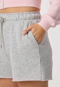 Hånd i lommen på grå shorts med elastisk talje, stylet med en lyserød langærmet crop top mod en ensfarvet baggrund.