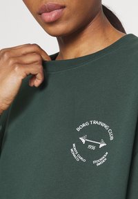 Sweat-shirt vert foncé avec un col côtelé. Présente un logo graphique blanc "BORG TRAINING CLUB" et un design de barre d'haltères.