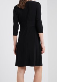 Musta mekko, jossa on muotoiltu vyötärö, kolmannen neljänneksen hihat ja polvimittainen hame. Sujuva pinta ja hienovarainen ompelu yksityiskohtana vyötäröllä.