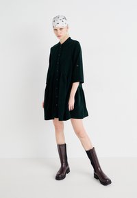 Robe en velours côtelé vert foncé avec fermeture à boutons, ourlet évasé, manches trois quarts. Portée avec des bottes en cuir marron hautes. Foulard blanc à motifs.