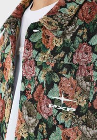 Denimjakke med blomsterprint og tekstureret stof, der har dominerende nuancer af rose og grønt, metalknapper og et broderet logo på brystet.