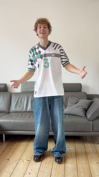 Balta džersija ar zaļiem akcentiem un ģeometriskām formām, uz kuras redzams numurs 3. Kombinēta ar vaļīgām zilām džinsa biksēm un melniem apaviem.