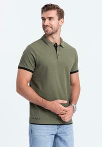 Polo shirt verde oliva con colletto e inserti neri sulle maniche, dotato di maniche corte e patta con tre bottoni, abbinato a jeans in denim chiaro.