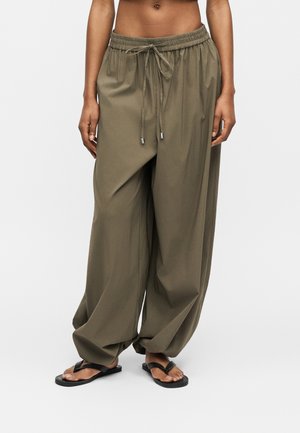 Femeie care poartă pantaloni largi verde măsliniu cu șnur și șlapi negri, stând pe un fundal simplu și deschis la culoare.