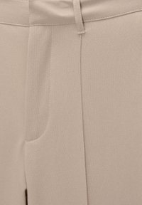 Gros plan de la taille et de l'avant d'un pantalon beige avec passants de ceinture visibles, fermeture éclair dissimulée et couture de poche latérale.