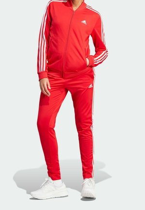 Personne portant un survêtement rouge Adidas avec des bandes blanches et des baskets blanches, debout avec une main dans la poche devant un fond uni.