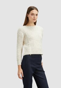 Off-white neulepaita, jossa on teksturoitu kuvio, pyöreä kaulus ja pitkät hihat, yhdistettynä tummiin farkkuihin, joissa on näkyvät saumat.