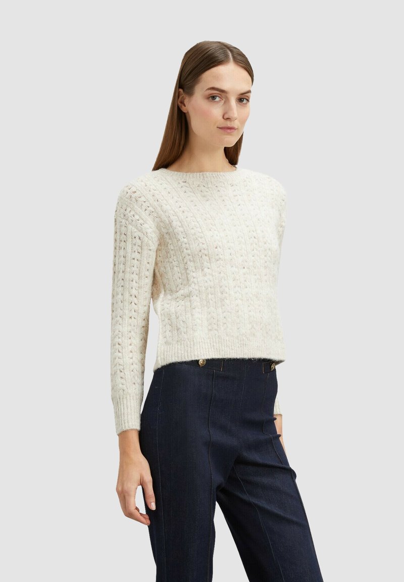 Off-white neulepaita, jossa on teksturoitu kuvio, pyöreä kaulus ja pitkät hihat, yhdistettynä tummiin farkkuihin, joissa on näkyvät saumat.