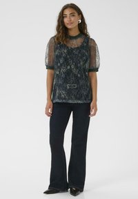 Frau mit welligem Haar, die ein dunkles, floral gemustertes, Spitzenoberteil mit Puffärmeln, enganliegende, dunkle Schlagjeans und spitze schwarze Schuhe vor einem weißen Hintergrund trägt.