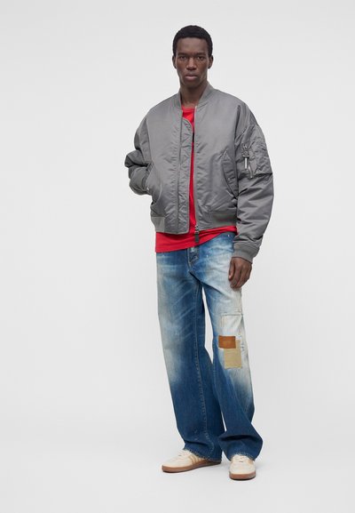Homme portant une veste bomber grise, une chemise rouge, un jean bleu large avec des patchs et des baskets blanches, debout devant un fond clair uni.