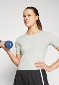 Lys grøn treningstrøye med korte ermer og rund hals, kombinert med mørke shorts med hvite sidedetaljer. En blå hantel holdes i den ene hånden.