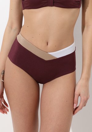 Donna che indossa slip bikini a vita alta color bordeaux con pannelli sovrapposti in vita beige e bianco, in piedi davanti a uno sfondo semplice.