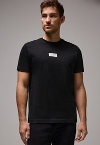 Svart bomull t-shirt med rund hals, korta ärmar och en rektangulär vit etikett med text placerad på bröstet.