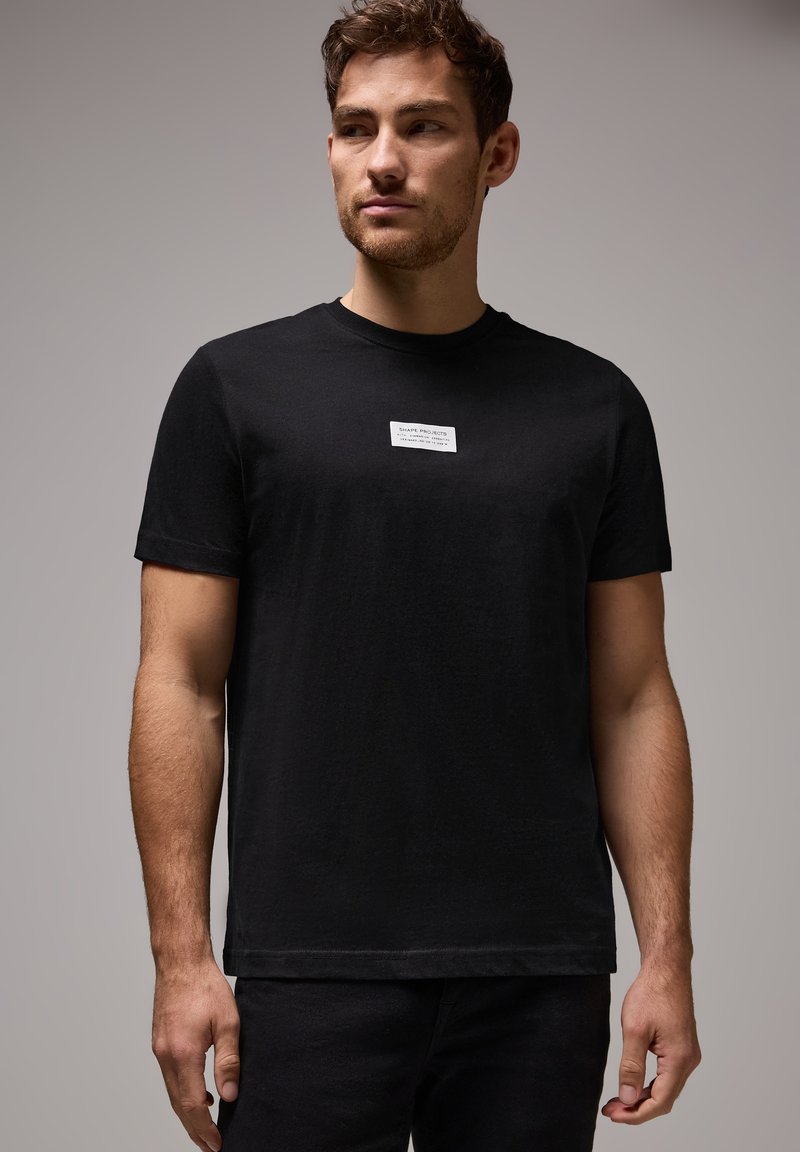 Svart bomull t-shirt med rund hals, korta ärmar och en rektangulär vit etikett med text placerad på bröstet.