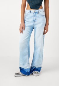 Vaaleansiniset, leveälahkeiset farkut, joissa on kulunut reuna ja tummansininen korostus lahkeensuussa. Yhdistetty mustavalkoisiin ruudullisiin slip-on-kenkiin.