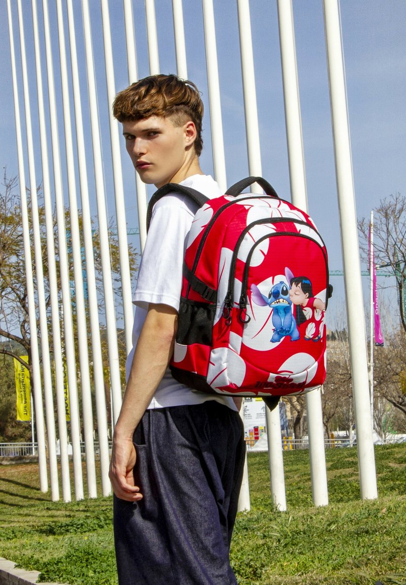 Mochila roja con un estampado de dibujos animados que presenta personajes azules y rosados, ribete negro y formas abstractas blancas. Cintas ajustables y bolsillos con cremallera.