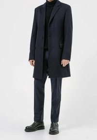 Abrigo de lana azul marino con cuello de solapa, tres botones, bolsillos laterales, combinado con un jersey de cuello alto de canalé y pantalones a medida, terminado con botas negras.