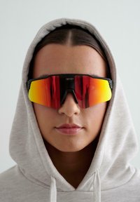 Persona con una sudadera ligera color gris y grandes gafas deportivas reflectantes rojas-anaranjadas, mirando hacia adelante contra un fondo liso.