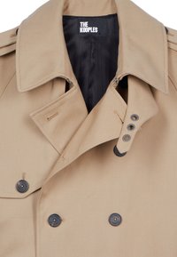 Trench coat beige con un cuello estructurado, diseño de doble botonadura, botones oscuros y cremalleras minimalistas. La tela parece suave y duradera.