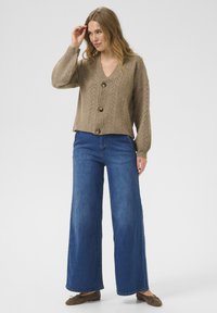 Cardigan en tricot beige avec un col en V, présentant un motif texturé et trois boutons. Associé à un jean bleu à jambes larges et des chaussures marron.