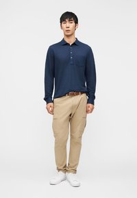 Man staand, gekleed in een marineblauw poloshirt met lange mouwen, beige cargobroek met riem en witte sneakers, tegen een lichte effen achtergrond.