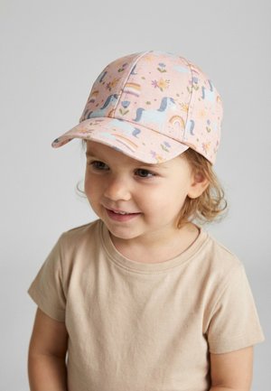 Junges Kind mit einer rosa Mütze mit blauen Pferden, Regenbögen und Blumen, lächelnd und leicht zur Seite blickend vor einem einfarbigen Hintergrund.