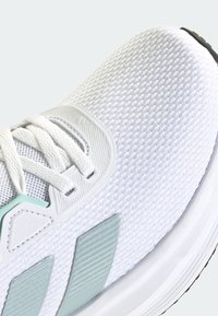 Gros plan d'une chaussure de sport blanche avec une tige en mesh texturé, des lacets blancs et des empiècements synthétiques bleu clair près de la semelle.