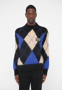 Man in een zwart, blauw en beige ruitpatroon sweater met een Versace Couture logo patch, gecombineerd met zwarte broek, staand tegen een witte achtergrond.