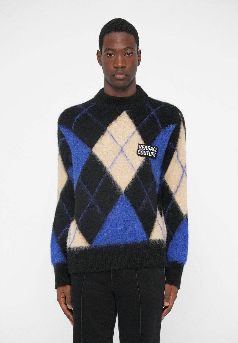 Man in een zwart, blauw en beige ruitpatroon sweater met een Versace Couture logo patch, gecombineerd met zwarte broek, staand tegen een witte achtergrond.