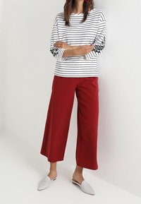 Witte gestreepte lange mouwen top met zwarte lijnen, gecombineerd met brede rode broeken. De look wordt afgemaakt met lichtgrijze instapschoenen.