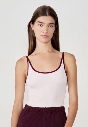 Joven mujer con cabello largo y marrón, vistiendo una camiseta sin mangas blanca con ribetes burdeos y pantalones burdeos a juego, frente a un fondo liso.