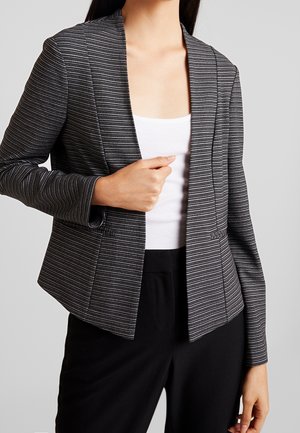 Femme portant un blazer rayé noir et gris par-dessus un haut blanc et un pantalon noir, tenant le blazer fermé d'une main.