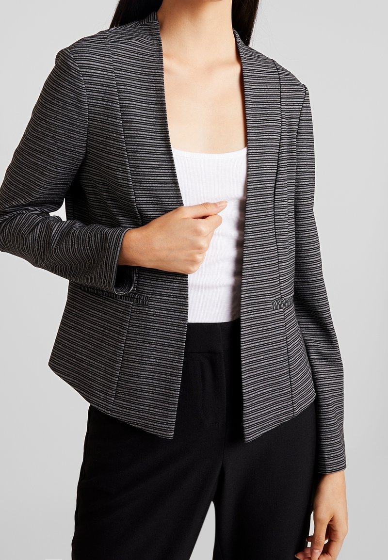 Femme portant un blazer rayé noir et gris par-dessus un haut blanc et un pantalon noir, tenant le blazer fermé d'une main.