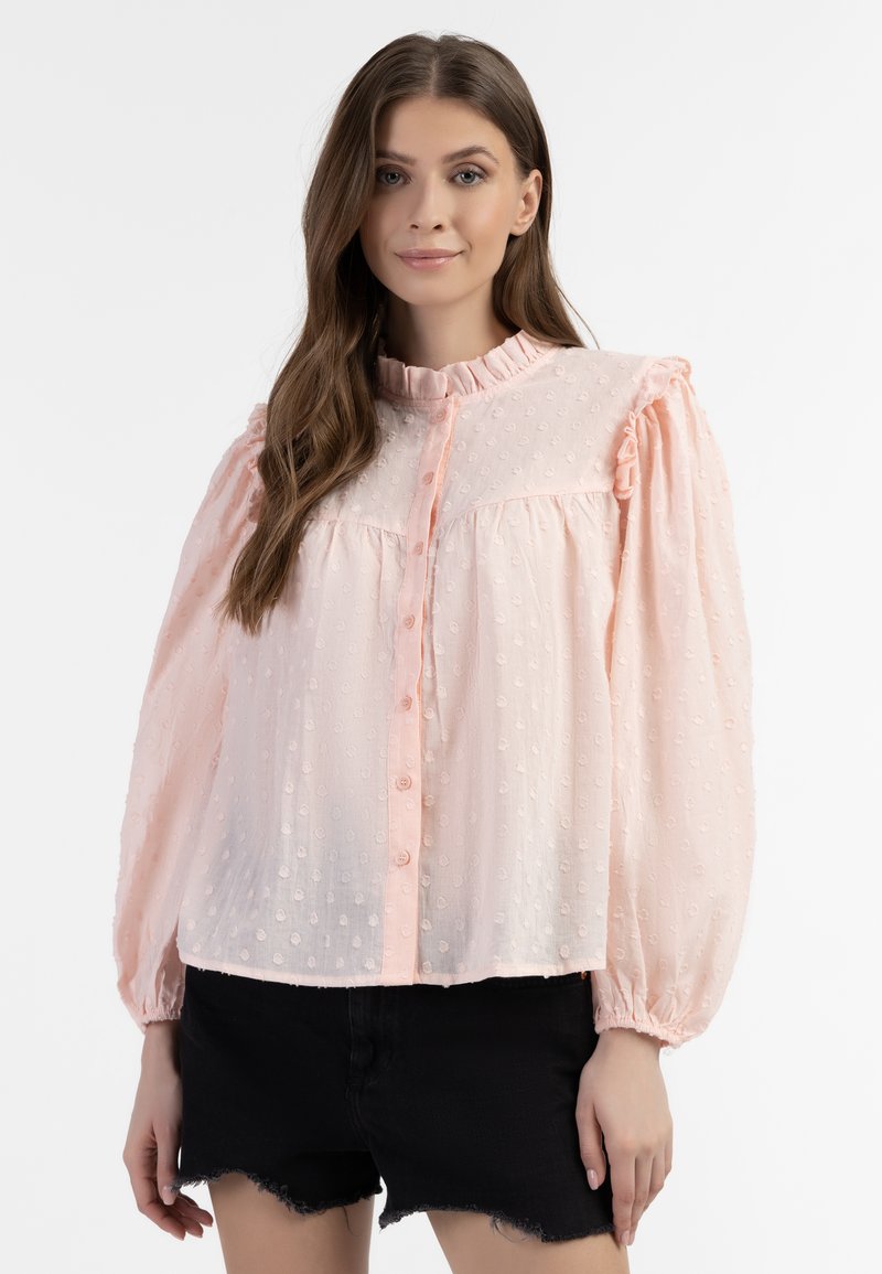 Lichtroze blouse van gestructureerde stof, met een ruches kraag, knoopsluiting en puffmouwen. Zwarte shorts worden eronder gedragen.
