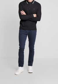Långärmad svart polo skjorta med vita krageakcent, tillsammans med marinblå slim-fit jeans och vita sneakers, som visar en avslappnad stil.