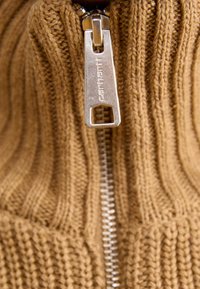 Brun stickad tröja med vertikal ribbning, med en silverdragkedja märkt "Carhartt." Närbild visar textur och detaljer.