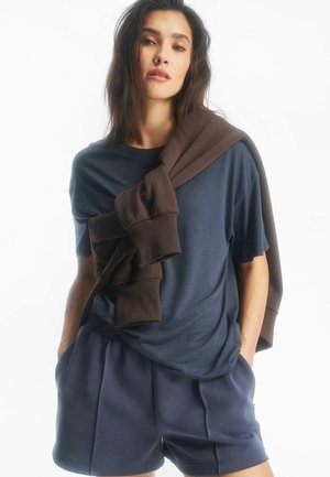 Kobieta w granatowej, krótko rękawowej koszulce i szortach, z ciemnobrązową bluzą przewieszoną przez ramiona, ręce w kieszeniach.