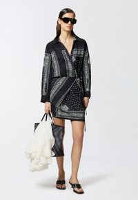 Abito avvolgente nero con intricati motivi e texture, dotato di maniche lunghe, colletto e lacci in vita. Abbinato a una borsa a tracolla nera e sandali.