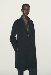 Cappotto in misto lana nero con revers, chiusura a singolo bottone, tasche laterali e silhouette ampia. Tessuto testurizzato, lunghezza fino al ginocchio.