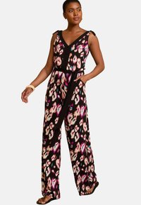 Ženska v črnem brezrokavnem kombinezonu s cvetličnimi motivi v roza, beli in modri barvi, širokimi hlačami in zavezanim pasom, stoji z eno roko v žepu.