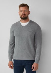 Grijze gebreide trui met een V-hals, ribgebreide mouwen en een klein logo-detail op de borst. Draagt over een wit T-shirt en gecombineerd met donkere broek.