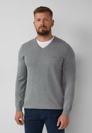 Grijze gebreide trui met een V-hals, ribgebreide mouwen en een klein logo-detail op de borst. Draagt over een wit T-shirt en gecombineerd met donkere broek.